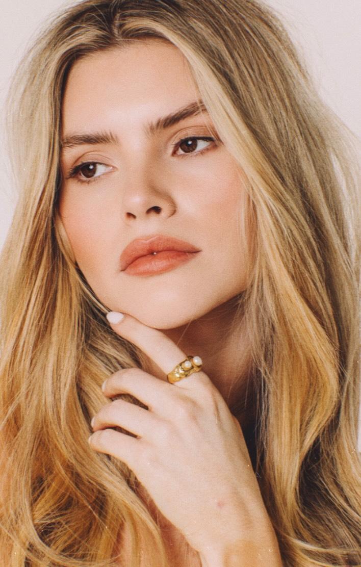Woman with long blonde hair and a gold ring on her finger against a plain background
