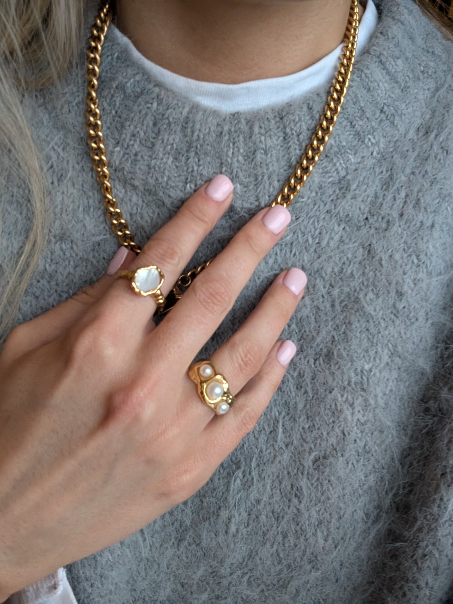 Close-up of a hand wearing gold rings with pearl-like stones, holding a gold chain necklace.