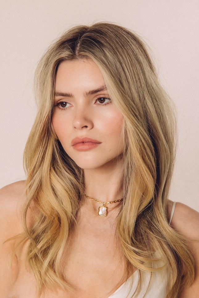 Woman with long blonde hair wearing a gold necklace against a light pink background